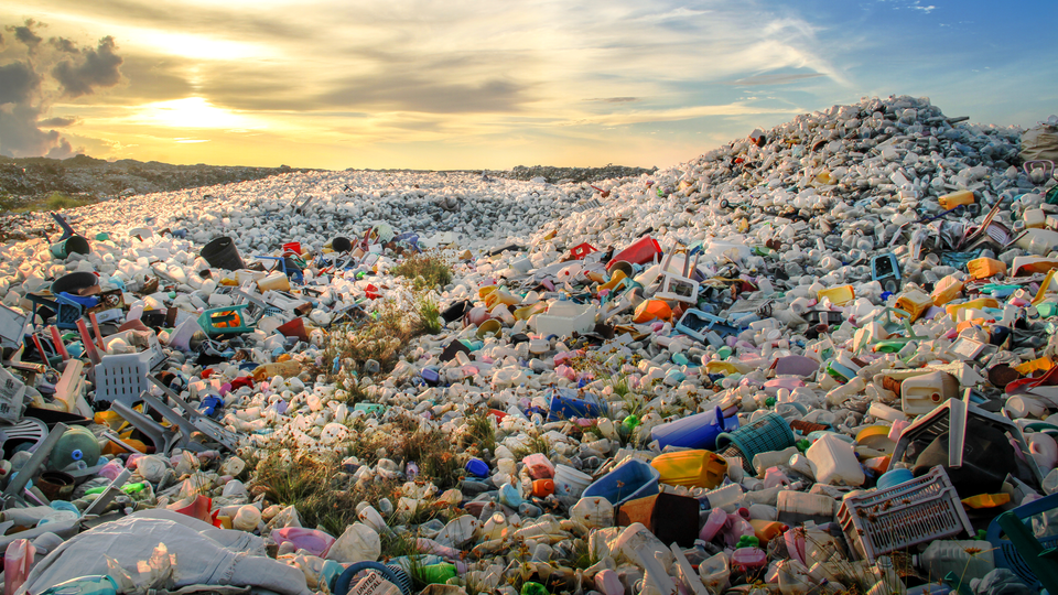 A landfill made up of small plastic items such as bottles and bowls, plastic stools ETC. One bigger pile in front of the landscape in the back, covered in plastic until the horizon which has a sunset. 